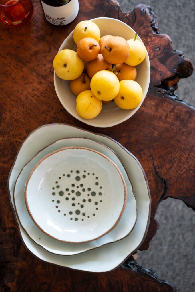This image contains 4 hand-crafted white ceramic bowls. Three are stacked and the fourth is overflowing with asian pears. The bowls sit on an old growth redwood slab coffee table and a bottle of wine and two glasses sit next to the bowls.